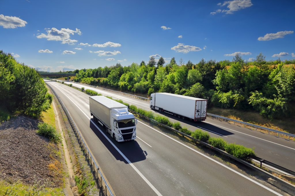 Two freight trucks driving on a divided highway surrounded by trees, symbolizing cross-border transportation and logistics movement
