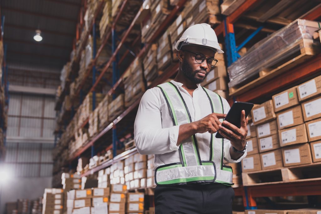 Warehouse worker in safety gear using a digital tablet among high shelves of inventory, illustrating modern fulfillment operations